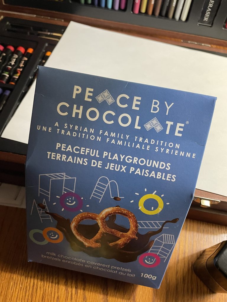 A pale blue package of Peaceful Playgrounds, milk chocolate covered pretzels, made by Peace By Chocolate. The package sites in front of an art kit and a blank sheet of paper.