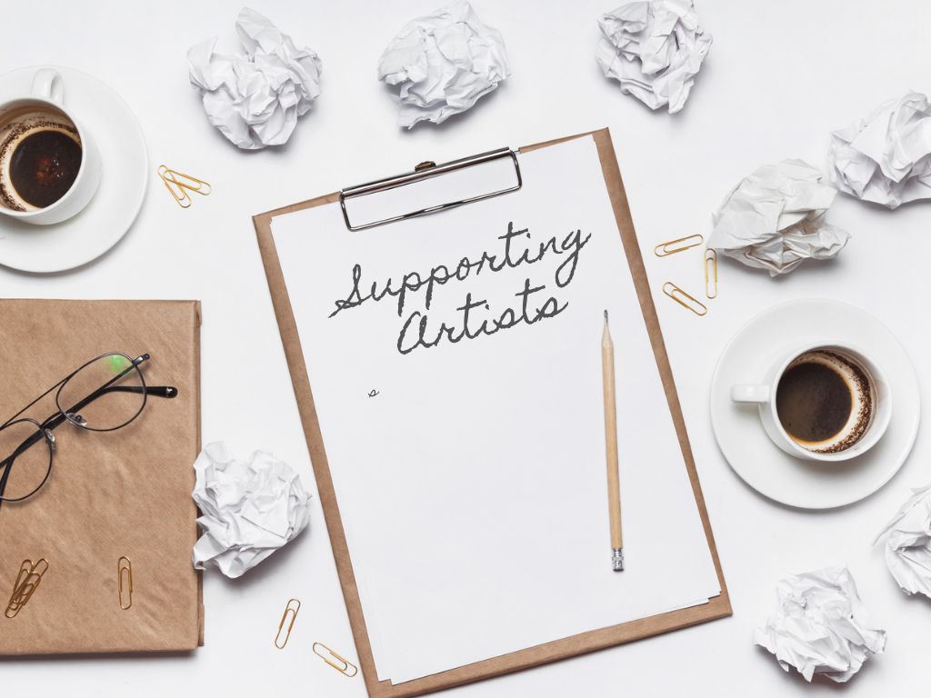 Support Artists on a clipboard sheet, while all around on the desk are scraps of paper, coffee cups, and the detritus of an artist's daily life.